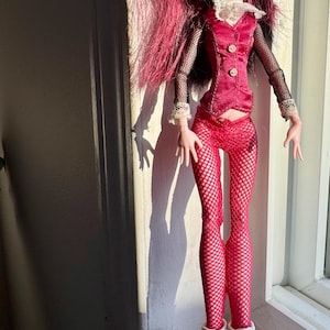 May include: A fashion doll with long, dark pink hair and pale skin. The doll wears a red vest, fishnet leggings, and white boots. The doll has a dramatic makeup look with dark lipstick and eyeshadow.