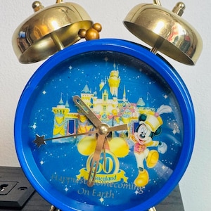 May include: A vintage blue alarm clock with a gold-colored frame and bells. The clock face features a Disney theme with Mickey Mouse, a castle, and the text "Happiest Homecoming On Earth". The clock has gold-colored legs and hands.