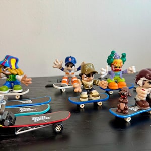 May include: A collection of miniature skateboarder figurines in various poses. The figures are on skateboards with black decks and colorful wheels. One skateboard is a yellow dragster. The figures are wearing hats and colorful outfits.