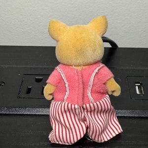 May include: A small, yellow-toned animal figurine wearing a pink outfit with white trim. The figurine's pants are red and white striped. The figure has small ears and is facing away from the camera.