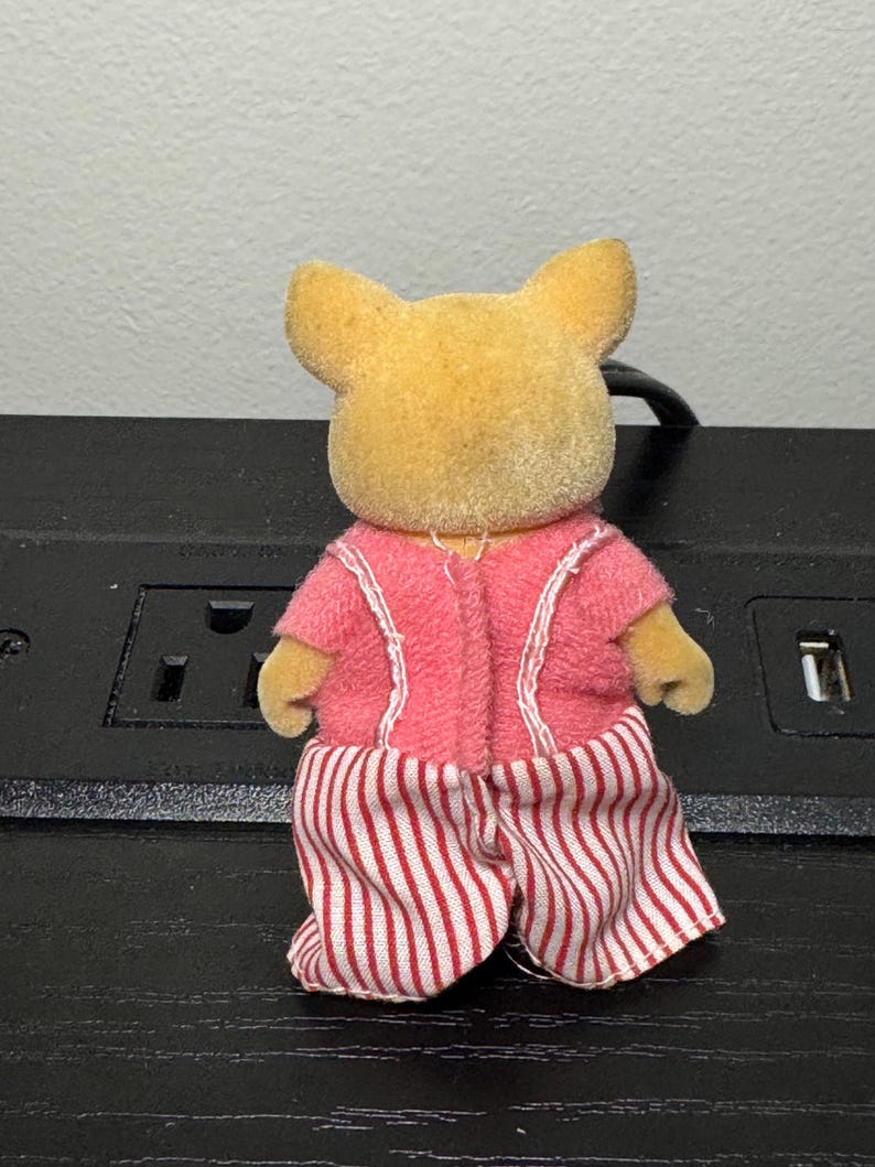 May include: A small, yellow-toned animal figurine wearing a pink outfit with white trim. The figurine's pants are red and white striped. The figure has small ears and is facing away from the camera.