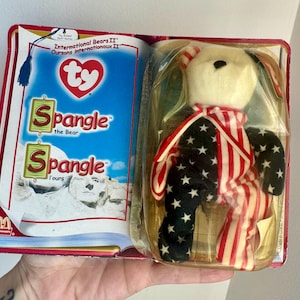 May include: A Spangle the Bear soft toy in its original packaging. The bear is white with red and white striped arms and a scarf, and black star-covered legs. The packaging features the text "Spangle the Bear" and "Spangle Tours".