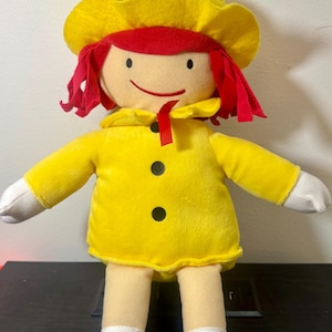 May include: A plush doll with a yellow hat and coat, red hair, and a smiling face. The doll has black button details and is wearing white socks and black shoes. The doll's arms are outstretched.