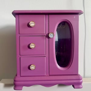 May include: A vibrant purple jewelry box with three drawers and a mirrored door. The box has decorative knobs with a circular design. The jewelry box is made of wood and has a scalloped base.