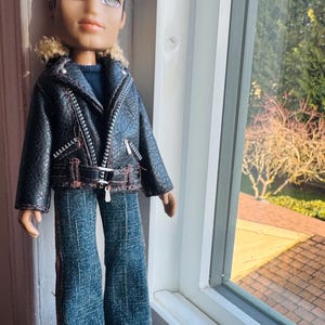 Könnte beinhalten: Eine Bratz-Puppe mit einer schwarzen Kunstlederjacke mit Kunstfellkragen, blauen Jeans und Schuhen mit Flammenmuster. Die Puppe hat dunkles Haar und steht in der Nähe eines Fensters.