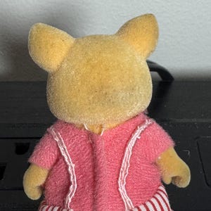 May include: A small, yellow-furred animal figurine wearing a pink jacket with white trim and red and white striped pants. The figurine has large, upright ears and small arms.