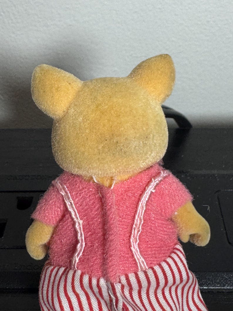 May include: A small, yellow-furred animal figurine wearing a pink jacket with white trim and red and white striped pants. The figurine has large, upright ears and small arms.