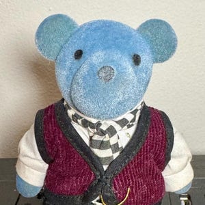 May include: A blue teddy bear wearing a burgundy corduroy vest, a black and white checkered scarf, and dark gray pants. The bear has blue ears and feet, and a gold-colored clasp on the vest.