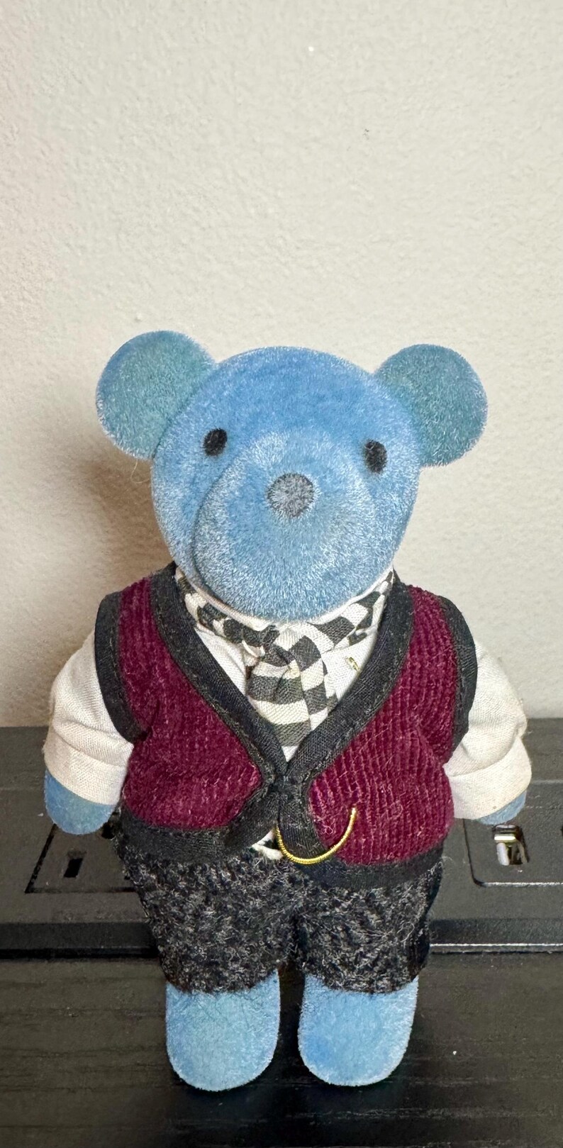 May include: A blue teddy bear wearing a burgundy corduroy vest, a black and white checkered scarf, and dark gray pants. The bear has blue ears and feet, and a gold-colored clasp on the vest.