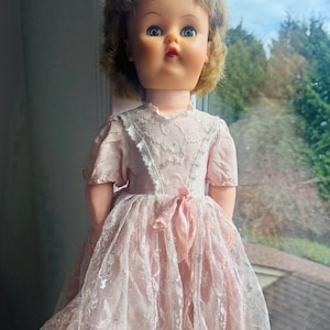 Puede incluir: Una muñeca vintage con cabello rubio que lleva un vestido de encaje rosa. La muñeca tiene ojos azules y una sonrisa pintada.