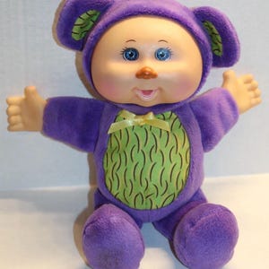 May include: A plush doll with a round face, blue eyes, and a small orange nose. The doll is wearing a purple outfit with a green patterned belly and a yellow bow. The outfit includes a hood with ears.