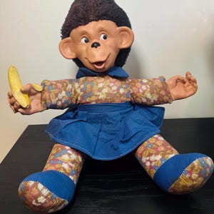 Può includere: Una bambola scimmia di peluche con viso in vinile stampato, capelli castani e grandi orecchie. La bambola indossa una gonna blu sopra una camicia a maniche lunghe con motivi floreali bianchi, rosa e verdi. Ha piedi in tessuto blu e tiene una banana.