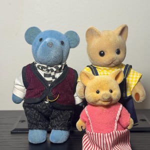 May include: Three miniature animal figurines: a blue bear in a vest and scarf, a tan bear in overalls, and a small pig in a pink dress with red and white striped pants. The toys are standing upright.