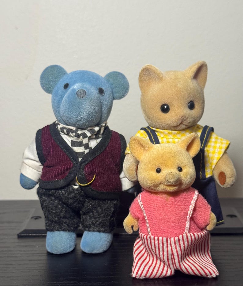 May include: Three miniature animal figurines: a blue bear in a vest and scarf, a tan bear in overalls, and a small pig in a pink dress with red and white striped pants. The toys are standing upright.