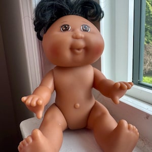 Op de afbeelding: Een vintage Cabbage Patch Kid pop met donker haar en bruine ogen. De pop heeft een gebruinde huidskleur en zit met uitgestrekte armen en benen. De pop is een klassiek verzamelobject.