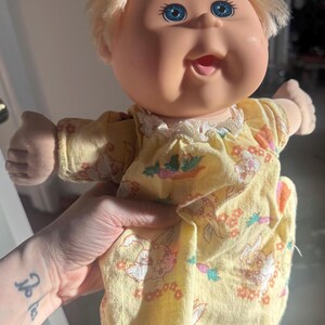 May include: A vintage doll with a round face, blue eyes, and blonde hair. The doll is wearing a yellow dress with a floral and bunny print, trimmed with white lace. The doll's arms are outstretched.