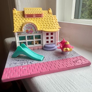 Op de afbeelding: Een plastic speelgoedwinkel-speelset met een geel dak, witte muren en een bord met de tekst TOYS. De set bevat een turquoise glijbaan, een roze vliegtuigje en een basis met een grijs gespikkeld gedeelte en een roze stoepgedeelte.