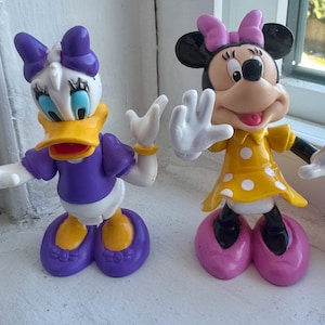 May include: Two plastic character figurines. On the left, a white duck with a yellow beak, purple shirt, purple bow, and purple shoes. On the right, a black mouse with a yellow dress featuring white polka dots, a pink bow, and pink shoes.