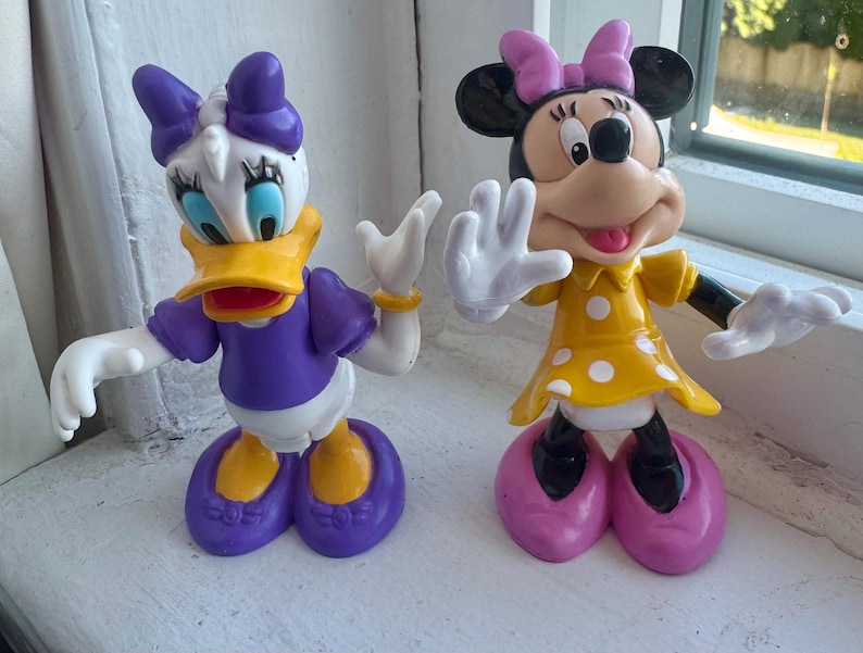 May include: Two plastic character figurines. On the left, a white duck with a yellow beak, purple shirt, purple bow, and purple shoes. On the right, a black mouse with a yellow dress featuring white polka dots, a pink bow, and pink shoes.