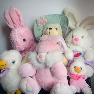 Puede incluir: Un conjunto de juguetes de peluche con temática de Pascua, que incluyen conejos rosas y blancos, un oso amarillo con sombrero y dos patitos blancos. Los conejos tienen orejas largas y narices rosas. Los patitos tienen picos amarillos y lazos.
