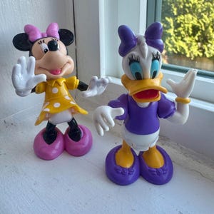 May include: Two plastic character figurines featuring Minnie Mouse and Daisy Duck. Minnie wears a yellow dress with white polka dots and a pink bow. Daisy Duck wears a purple top with a purple bow and yellow shoes. Both figures have white gloves.