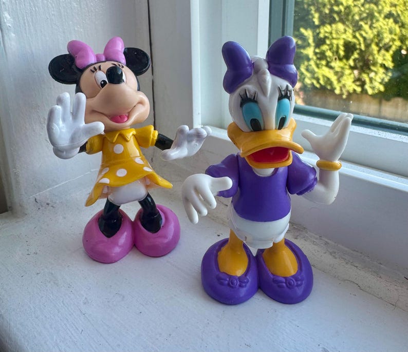 May include: Two plastic character figurines featuring Minnie Mouse and Daisy Duck. Minnie wears a yellow dress with white polka dots and a pink bow. Daisy Duck wears a purple top with a purple bow and yellow shoes. Both figures have white gloves.