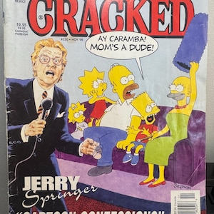 May include: A November 1998 edition of Cracked magazine with Jerry Springer on the front cover. The artwork shows Jerry Springer with a microphone, alongside the Simpsons family on a purple sofa. Text includes "Simpsons", "Small Soldiers", "Cracked", "Ay Caramba! Mom's a Dude!", "Jerry Springer", and "Cartoon Confessions".