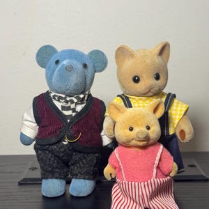 May include: Three miniature animal figurines. A blue bear figure wears a burgundy vest and checkered scarf. A tan cat figure wears a yellow checkered shirt and blue overalls. A tan pig figure wears a pink top and red and white striped pants.