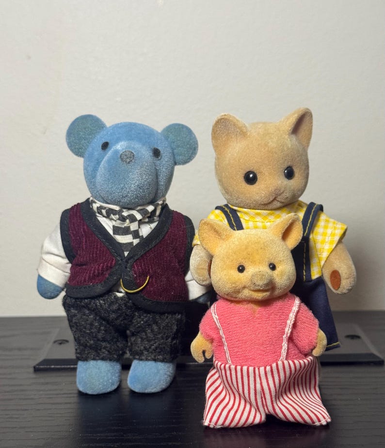 May include: Three miniature animal figurines. A blue bear figure wears a burgundy vest and checkered scarf. A tan cat figure wears a yellow checkered shirt and blue overalls. A tan pig figure wears a pink top and red and white striped pants.
