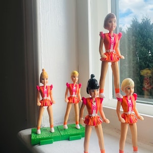 May include: A group of vintage cheerleader dolls. Each doll is wearing a vibrant orange and pink uniform with a "B" on the chest. The dolls have different hairstyles and are positioned on a green base, creating a nostalgic and playful display.