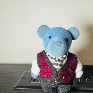 May include: A blue teddy bear figurine wearing a burgundy vest, a black and white striped scarf, and gray patterned pants. The bear has black eyes and a black nose. The figurine is standing on a black surface.