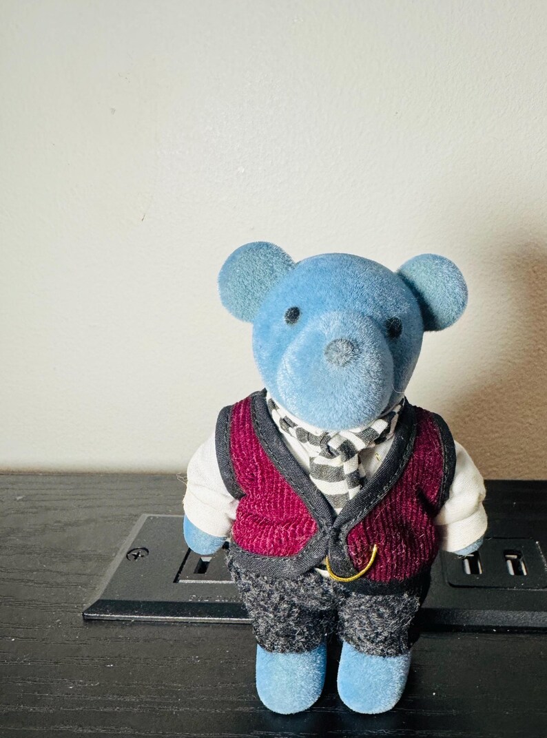 May include: A blue teddy bear figurine wearing a burgundy vest, a black and white striped scarf, and gray patterned pants. The bear has black eyes and a black nose. The figurine is standing on a black surface.