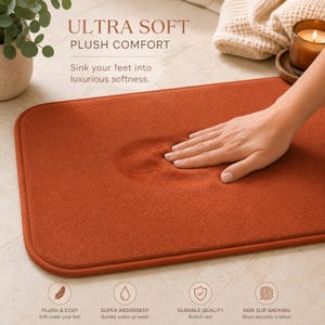 May include: An orange rectangular bath mat with rounded corners. The mat is plush and soft, with the text "ULTRA SOFT PLUSH COMFORT" at the top. A hand presses into the mat, showing its softness. The mat is described as plush, absorbent, durable, and non-slip.
