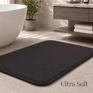 May include: A rectangular, dark gray bath mat with rounded corners. The mat is placed on a tiled floor in a bathroom setting. The words "Ultra Soft" are printed in white on the bottom right corner.