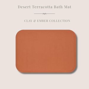 Puede incluir: Una alfombra de baño rectangular de terracota desértica, de color marrón anaranjado cálido. La alfombra tiene esquinas redondeadas. El texto "Desert Terracotta Bath Mat" y "Clay & Ember Collection" se muestra encima de la alfombra.