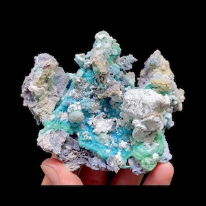 May include: A close-up of a mineral specimen with a complex, layered structure. The mineral features a vibrant turquoise hue, contrasted by patches of white, gray, and pale green. The specimen is held against a black background, highlighting its intricate details.