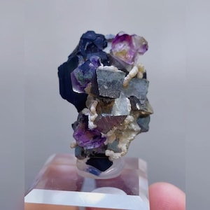 May include: A cluster of natural fluorite crystals. The crystals display a mix of colors, including deep purple, clear, and gray. The fluorite is mounted on a clear, rectangular base. The overall appearance is of a raw, unpolished mineral specimen.