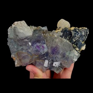 May include: A cluster of fluorite crystals with a mix of purple, clear, and gray hues. The mineral specimen has a crystalline structure with a dark background. The fluorite is a natural mineral.