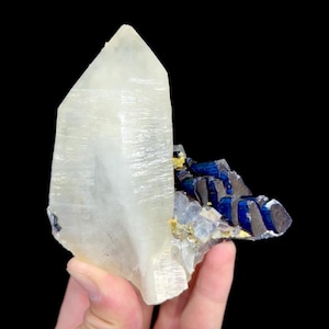 May include: A large, clear quartz crystal with a pointed tip, next to a cluster of dark blue and gray mineral formations. The crystal is held against a black background, showcasing its natural beauty and texture.