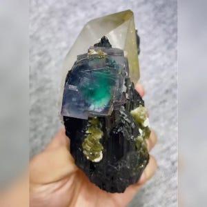 May include: A natural mineral specimen featuring a large, clear quartz crystal with a yellow hue at the top. The base is a cluster of black tourmaline crystals, with a vibrant green and blue fluorite cube and small green crystals.