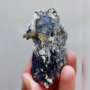 May include: A natural fluorite mineral specimen. The dark blue and gray stone features a square, translucent blue crystal. The specimen has a rough, textured surface with small, light-colored crystal formations.