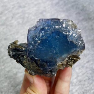 May include: A vibrant blue fluorite crystal cluster with a layered, geometric structure. The crystal displays a deep, saturated blue hue in its center, transitioning to lighter shades towards the edges. The fluorite is set on a dark, mineral-rich matrix.