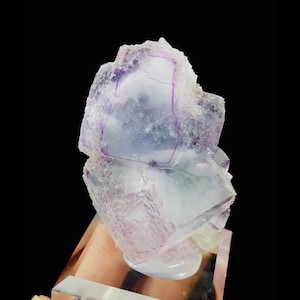 May include: A translucent fluorite crystal specimen with a layered structure. The crystal features shades of purple, lavender, and white, with a crystalline texture. It is mounted on a clear, rectangular base against a black background.