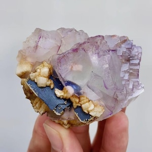 May include: A natural fluorite crystal cluster with a mix of colors. The crystal features translucent purple and white sections, with accents of blue and tan. The fluorite has a cubic structure and is a unique mineral specimen.