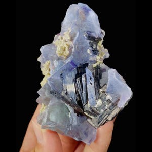 May include: A cluster of blue and purple fluorite crystals with dark, metallic inclusions and patches of light yellow minerals. The fluorite has a layered appearance, with sharp edges and a crystalline structure. The background is black.