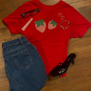 Peut inclure: Un t-shirt rouge avec deux appliqués de fraises, associé à un jean bleu, des lunettes à monture noire, un tube de brillant à lèvres, des boucles d'oreilles créoles dorées, un bracelet et des sandales à talons noires. Sur une surface en bois.