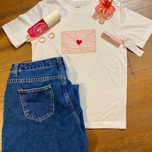 May include: White t-shirt with a pink and red embroidered envelope with a heart on the front. The shirt is laying on a wooden surface with a pair of gold earrings, a pink bottle of hand sanitizer, a pink lip gloss, and a pair of blue jeans.
