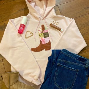 May include: A pink hoodie with a patchwork boot design on the front. The hoodie is laid out on a wooden surface with a pair of blue jeans, a pair of brown boots, a pink spray bottle, a gold chain necklace, and a tube of lip balm.