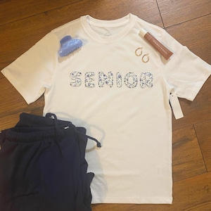 May include: A white t-shirt with the word "SENIOR" in floral print, paired with navy blue sweatpants. Accessories include a blue hair clip, gold hoop earrings, and a tube of lip gloss.