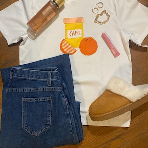 May include: White t-shirt featuring a graphic of a jar of jam and two orange slices. The shirt is styled with blue jeans, brown fluffy slippers, a bottle of body spray, a pink lip gloss, and gold jewellery.